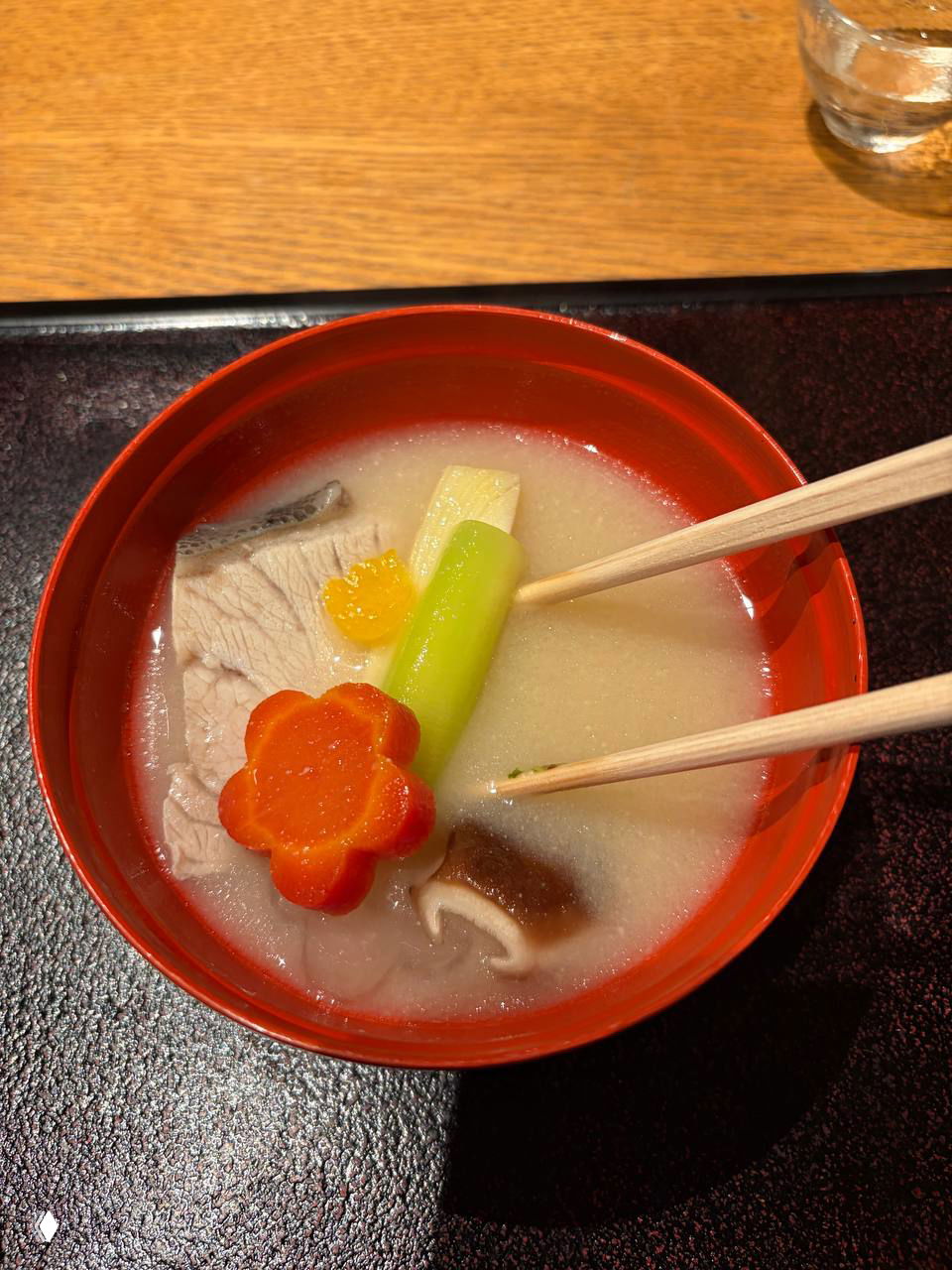 Красная миска с прозрачным бульоном, кусками рыбы, декоративной морковкой в форме цветка и овощным стеблем — традиционный вкус.