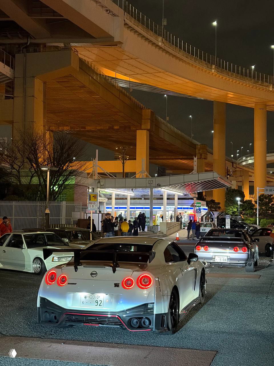 Ночной паркинг у развязки: серебристый Nissan GT‑R и другие тюнингованные машины на фоне высоких эстакад и уличного освещения, тусовка JDM.
