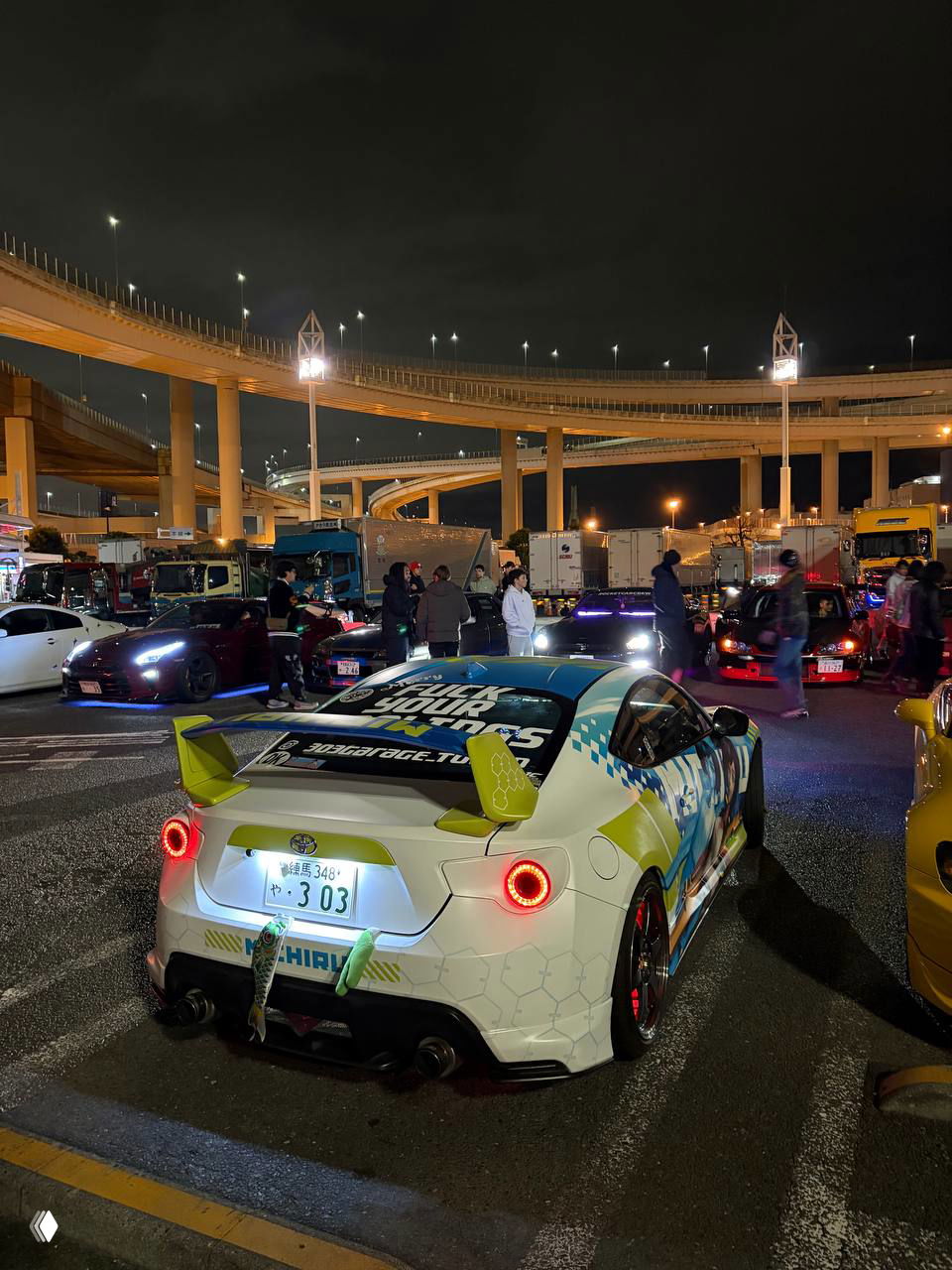 Широкий план парковки у дорожной развязки: люди группами, множество разнообразных автомобилей и яркое освещение городской ночной сцены тюнинг‑встречи.