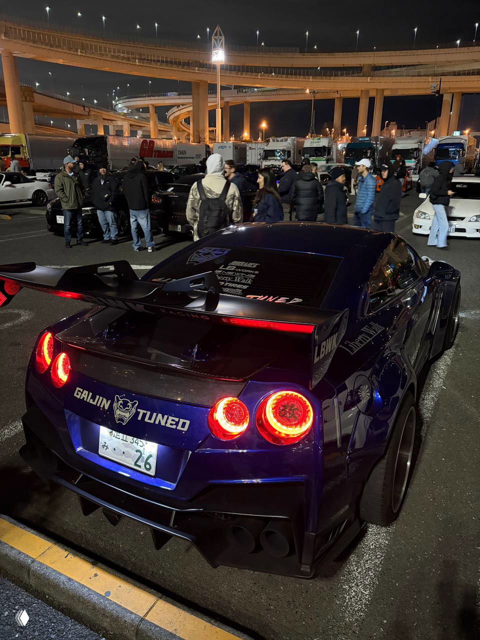 Белая или светлая машина с крупным антикрылом и яркими декоративными элементами, заметны светящиеся фонари и гоночный стиль в задней части кузова.