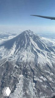 Вид из самолёта на высокий заснеженный вулкан Камчатки: конический пик, снежные склоны и часть крыла в кадре.