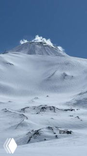 Заснеженный склон действующего вулкана с видимыми лавовыми структурами под снегом и шлейфом пара на вершине в солнечную погоду.