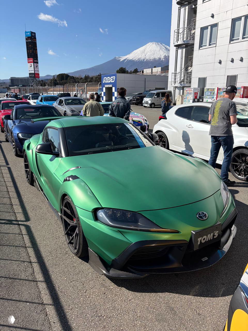 Общий план пит‑лейна: ряд спорткаров с зелёной Supra на переднем плане и заснеженной горой Фудзи на дальнем плане, люди у машин.