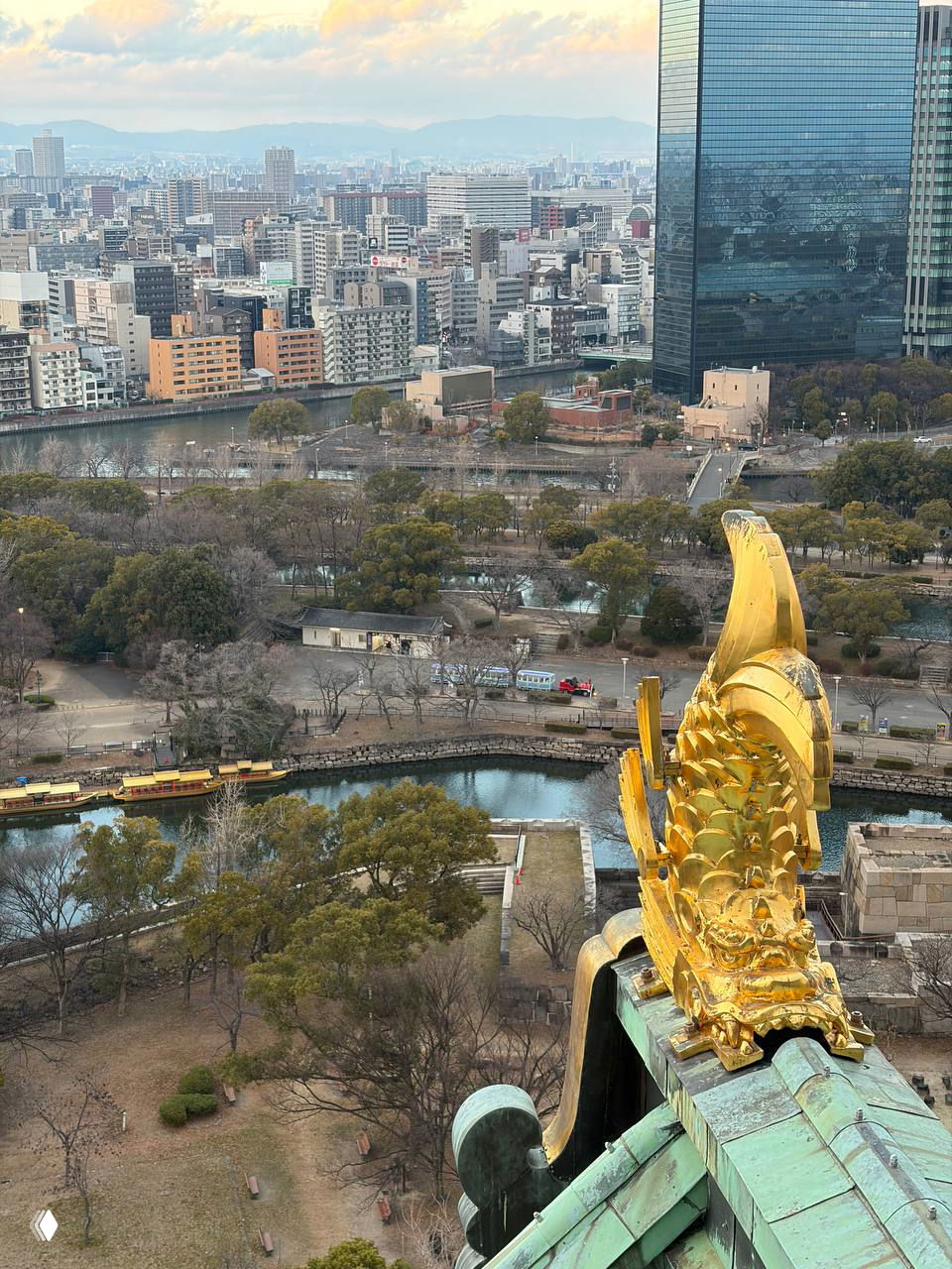 Панорамный городской пейзаж Осаки с видом на реку и парк, на переднем плане декоративная золотая фигурка в архитектуре крыши замка.